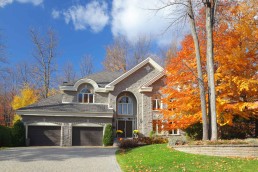 Large luxurious two-story residential house during autumn framed by tall trees with golden orange foliage partially masking one side of the home.