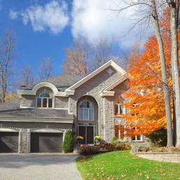 Large luxurious two-story residential house during autumn framed by tall trees with golden orange foliage partially masking one side of the home.